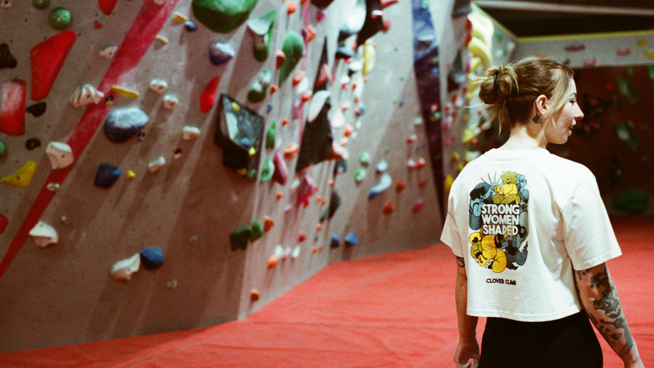 Woman standing in front of a colorful climbing wall wearing a t-shirt with a graphic design.